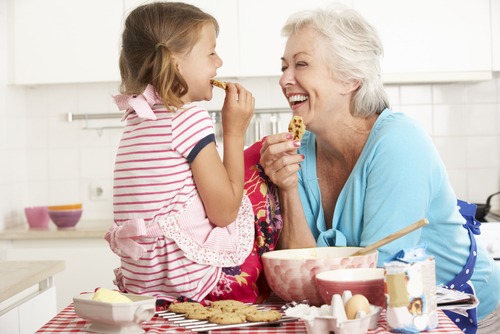 Image of a smiling grandparent seated beside their grandchild, both enjoying cookies together. The grandparent wears dentures, contributing to their joyful expression as they interact with their grandchild.